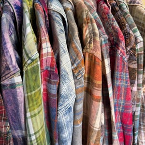 May include: A collection of long-sleeve button-down shirts hanging side by side, featuring various plaid and checkered patterns in colors including purple, green, red, blue, orange, and brown, with a distressed or bleached fabric texture.