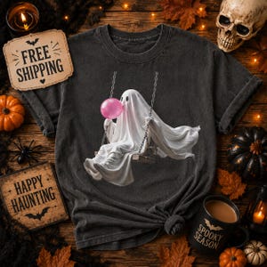 Puede incluir: Una camiseta gris carbón con un gráfico de un fantasma blanco sentado en un columpio y soplando una burbuja de chicle rosa. El fantasma está cubierto con una sábana blanca y el diseño está centrado en el pecho de la prenda de cuello redondo.