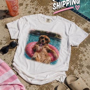 Op de afbeelding: Een wit t-shirt met korte mouwen en een grafische print van een golden retriever met een zwarte zonnebril, drijvend in een zwembad op een roze donutvormige zwemband, terwijl het dier een doorzichtige beker met een roze rietje vasthoudt.