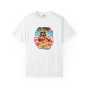 Könnte beinhalten: Ein weißes T-Shirt mit kurzen Ärmeln und einem Grafikdruck einer orangefarbenen Katze mit schwarzer Sonnenbrille, die in einem blauen Swimmingpool auf einem rosa-weiß gestreiften Schwimmring treibt und ein rotes Getränk mit Strohhalm hält.