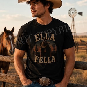 Puede incluir: Una camiseta negra de manga corta con un estampado gráfico con el texto ELLA FELLA, la silueta de una persona con sombrero de vaquero, un cactus y un molino de viento. La camiseta se lleva con vaqueros azules y una hebilla de cinturón metálica.
