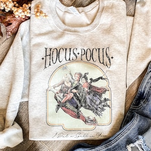 May include: A light gray crewneck sweatshirt featuring a graphic print of three witches flying on a broomstick. The text HOCUS POCUS is printed above the illustration, and the phrase I Put a Spell on You is printed below in a cursive script font.
