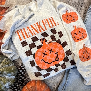 May include: A light gray sweatshirt with the word "THANKFUL" in orange block letters. The shirt features a checkered pumpkin graphic and three smaller pumpkin graphics on the sleeve. The sweatshirt is part of a fall-themed collection.