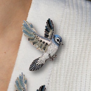 Può includere: Due spille a forma di merlo blu con ali e corpi in perle bianche e blu. Le spille sono su un maglione bianco.