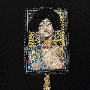 Può includere: Spilla ricamata con un ritratto di donna con afro scuro. Il ritratto è incorniciato in nero e oro, con un pendaglio a catena dorata. Il volto della donna è dettagliato con piccole perline di vari colori.