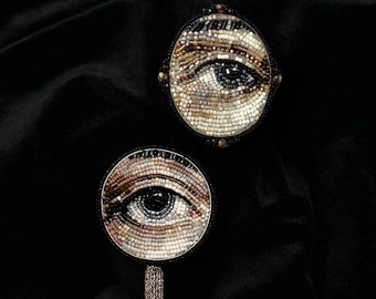 Peut inclure: Deux broches rondes en perles avec un motif d'œil. Les broches sont sur un fond de tissu noir. Une broche a un pompon en chaîne argentée.
