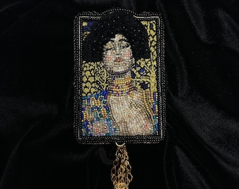 May include: A beaded brooch featuring a portrait of a woman with dark hair and a gold necklace. The brooch is framed in black and has a gold chain tassel.