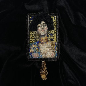 Può includere: Un broche di perline con il ritratto di una donna con i capelli scuri e una collana d'oro. Il broche è incorniciato in nero e ha un fiocco di catena dorata.