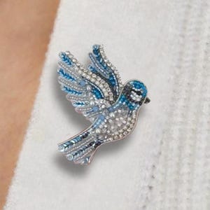 May include: A jeweled bird brooch with blue and silver-colored crystals. The bird is in flight, with its wings spread. The brooch is pinned to a white sweater.