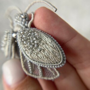 May include: A detailed, handmade bee brooch. The wings are a light gray, with intricate beadwork and delicate embroidery. The body is covered in small, silver beads, and the antennae are made of wire. This unique accessory adds a touch of nature-inspired elegance.