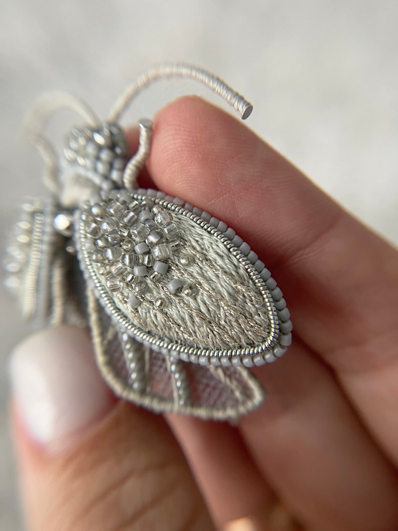 May include: A detailed, handmade bee brooch. The wings are a light gray, with intricate beadwork and delicate embroidery. The body is covered in small, silver beads, and the antennae are made of wire. This unique accessory adds a touch of nature-inspired elegance.