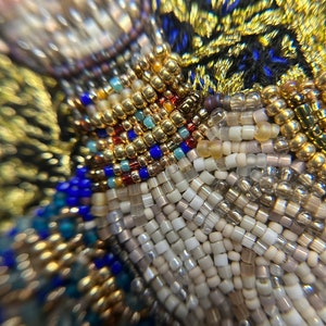 May include: Close-up of a beaded fabric with a variety of colors, including gold, white, blue, and red. The beads are arranged in a pattern that creates a textured surface.