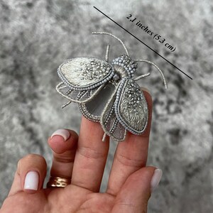 May include: A silver-colored, beaded insect brooch, measuring 2.1 inches (5.3 cm) in length. The brooch features intricate detailing with a combination of beads and embroidery, creating a delicate and ornate design. The wings and body are embellished with small, shiny beads.