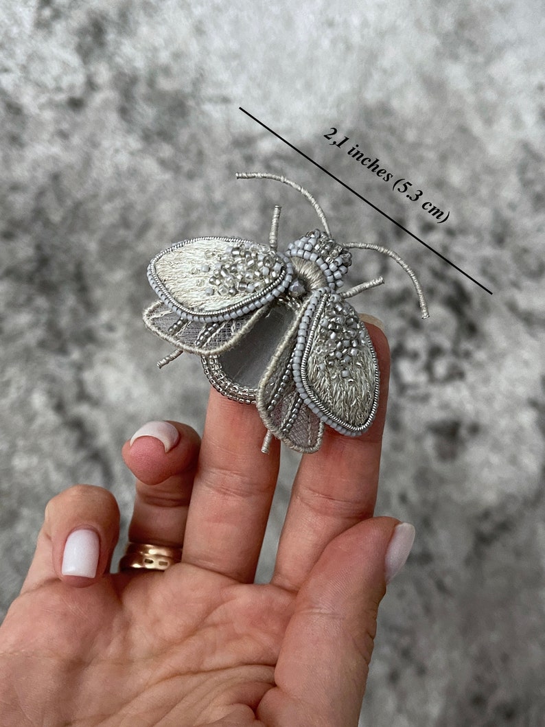 May include: A silver-colored, beaded insect brooch, measuring 2.1 inches (5.3 cm) in length. The brooch features intricate detailing with a combination of beads and embroidery, creating a delicate and ornate design. The wings and body are embellished with small, shiny beads.