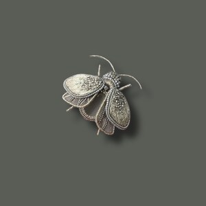 May include: A detailed silver-toned insect brooch. The brooch features intricate beadwork and textured wings. The insect has a segmented body and delicate antennae. The brooch is set against a solid gray background, highlighting its fine craftsmanship.