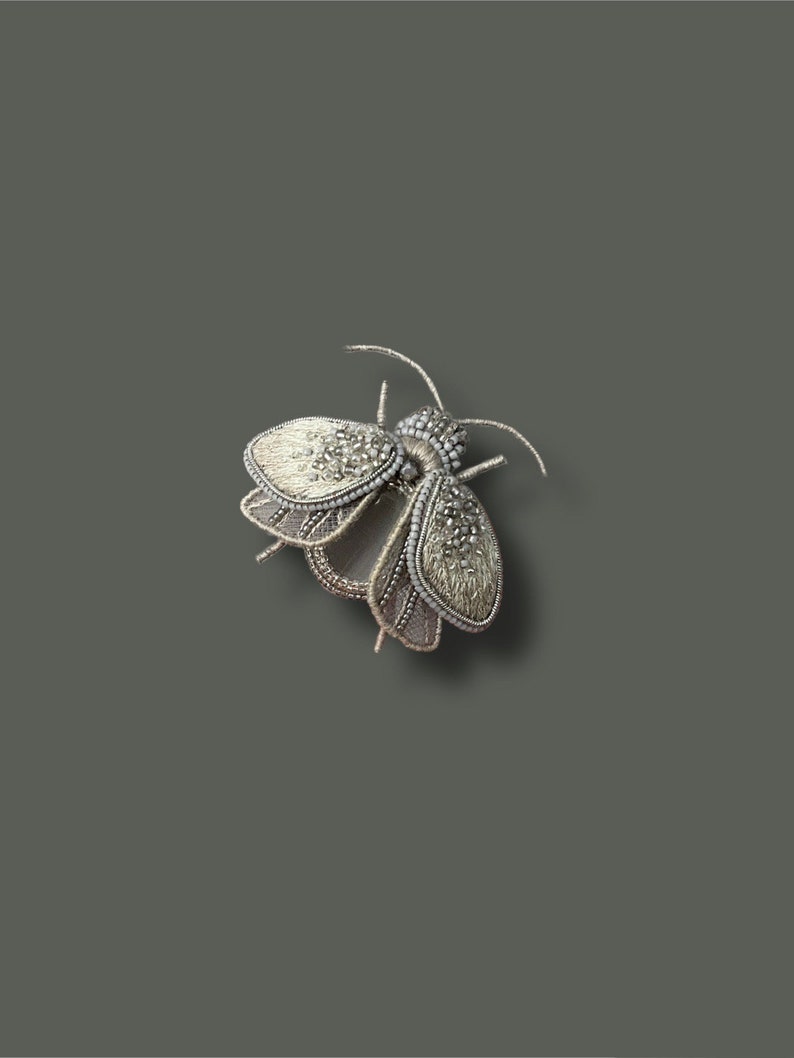 May include: A detailed silver-toned insect brooch. The brooch features intricate beadwork and textured wings. The insect has a segmented body and delicate antennae. The brooch is set against a solid gray background, highlighting its fine craftsmanship.