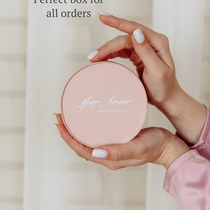May include: A round, light pink box with the words "Your Amour" and "Marina Mankevych" in elegant script. The box is held in two hands with white nail polish. The text "Perfect box for all orders" is in the background.
