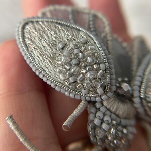 May include: A close-up of a handmade, silver-colored brooch in the shape of a stylized insect. The brooch features intricate beadwork and embroidery, with a delicate, textured appearance. The wings and body are adorned with small, clear and silver beads.