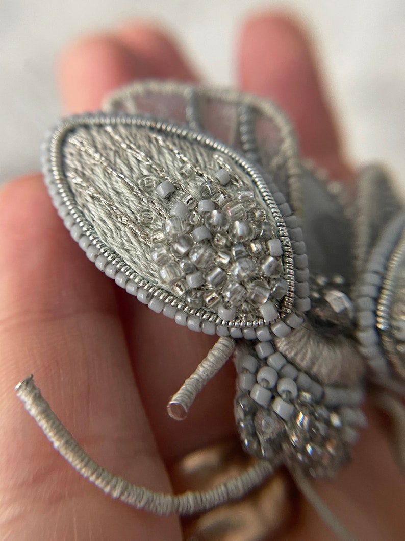 May include: A close-up of a handmade, silver-colored brooch in the shape of a stylized insect. The brooch features intricate beadwork and embroidery, with a delicate, textured appearance. The wings and body are adorned with small, clear and silver beads.