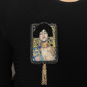 May include: A beaded brooch featuring a portrait of a woman with a gold chain tassel. The portrait is framed in a gold and black beaded border.