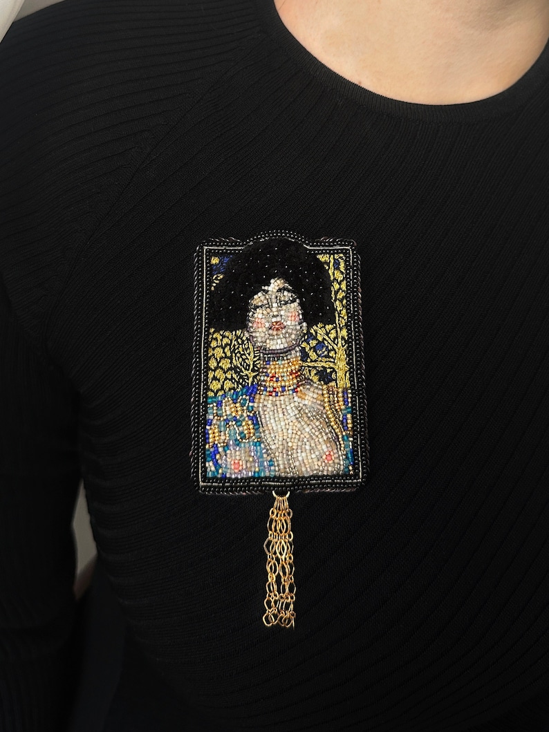 May include: A beaded brooch featuring a portrait of a woman with a gold chain tassel. The portrait is framed in a gold and black beaded border.