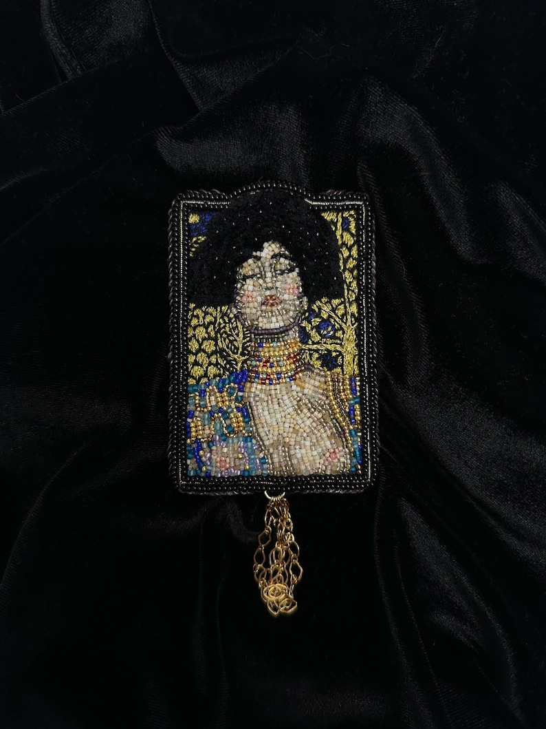 May include: A beaded brooch featuring a portrait of a woman with dark hair and a gold necklace. The brooch is framed in black and has a gold chain tassel.