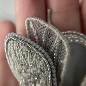 May include: Close-up of a gray, beaded butterfly brooch. The brooch features intricate embroidery and delicate beadwork, with a silver wire outline. The wings have a lacy, sheer texture, and the overall design is elegant and detailed.
