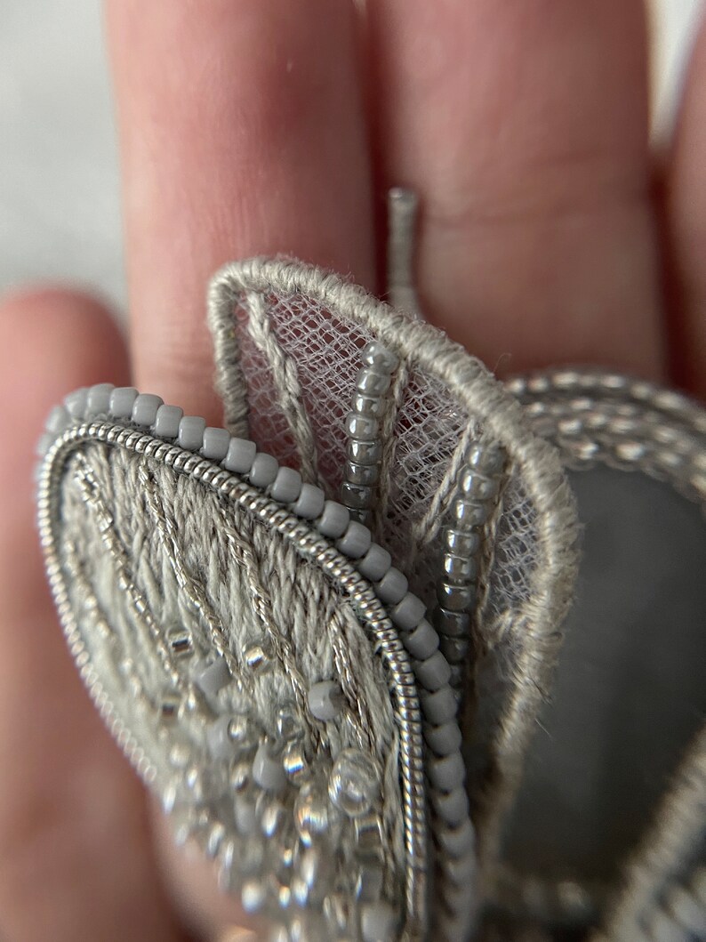 May include: Close-up of a gray, beaded butterfly brooch. The brooch features intricate embroidery and delicate beadwork, with a silver wire outline. The wings have a lacy, sheer texture, and the overall design is elegant and detailed.