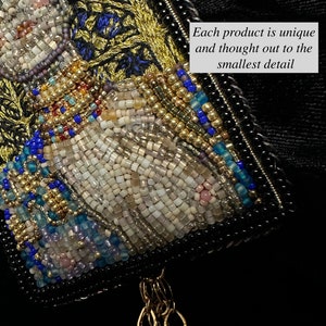 May include: A close-up of a beaded artwork featuring a white figure with gold and blue accents. The artwork is framed with black beads and has a gold chain tassel.
