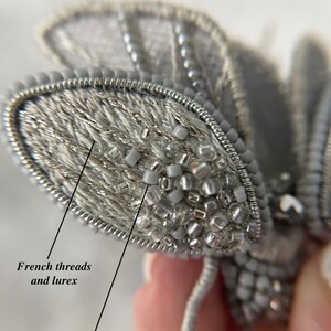 May include: A silver-plated brooch pin in the shape of a moth. The moth's wings are made with French threads and lurex, and the body is adorned with Japanese beads. The brooch is a detailed piece of jewelry.