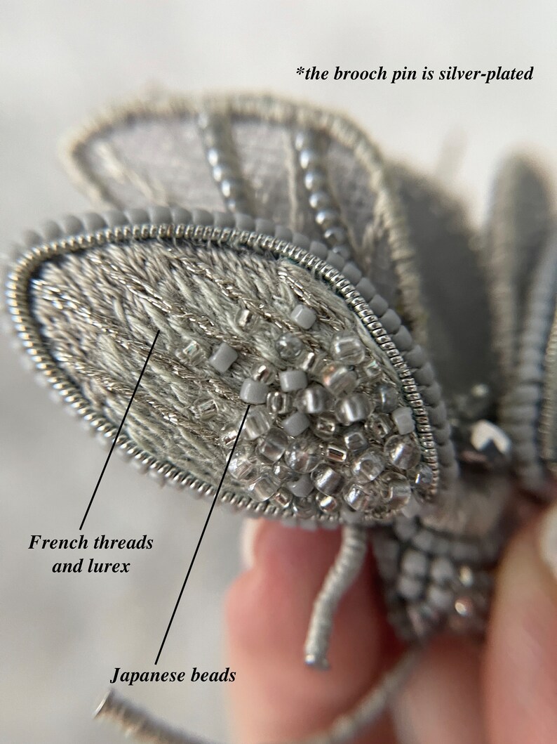 May include: A silver-plated brooch pin in the shape of a moth. The moth's wings are made with French threads and lurex, and the body is adorned with Japanese beads. The brooch is a detailed piece of jewelry.
