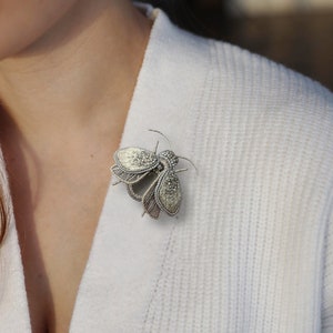 May include: A silver-toned insect brooch with intricate detailing, pinned to a white sweater. The brooch features a detailed design with textured wings and a beaded body, creating a delicate and elegant accessory. This insect-themed brooch is a unique piece of jewelry.