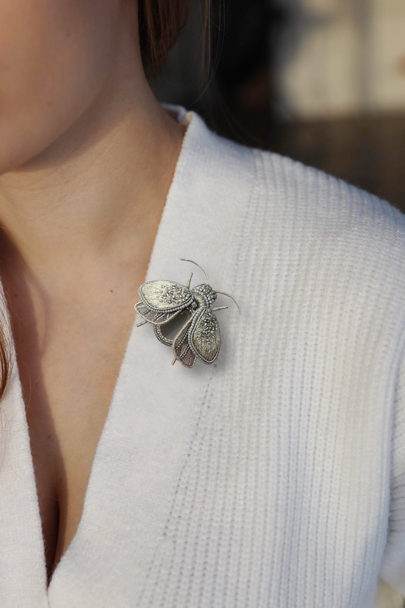 May include: A silver-toned insect brooch with intricate detailing, pinned to a white sweater. The brooch features a detailed design with textured wings and a beaded body, creating a delicate and elegant accessory. This insect-themed brooch is a unique piece of jewelry.
