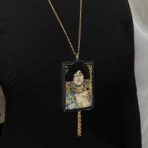 May include: A gold chain necklace with a beaded pendant featuring a portrait of a woman with dark hair and a gold crown. The pendant is framed in black and has a gold tassel.