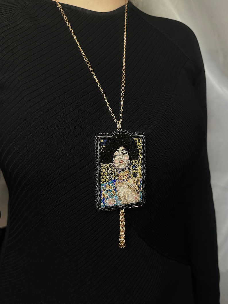 May include: A gold chain necklace with a beaded pendant featuring a portrait of a woman with dark hair and a gold crown. The pendant is framed in black and has a gold tassel.