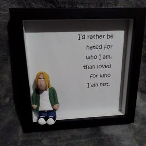 May include: A framed artwork with a clay figure and text. The figure, with blonde hair and a green jacket, is next to the text "I'd rather be hated for who I am, than loved for who I am not." The frame is black with a white background.