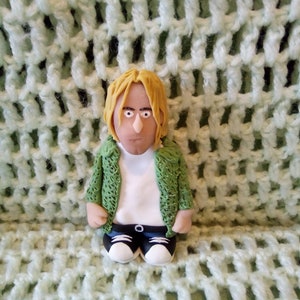 May include: A handmade figurine of a person with blonde hair, wearing a green jacket over a white shirt, black trousers, and white trainers. The figurine is set against a light green, textured background.