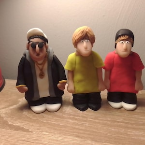 May include: Three handmade figurines of people. The figure on the left is wearing a black jacket, a white shirt, and a gold chain. The figure in the middle is wearing a yellow shirt and black trousers. The figure on the right is wearing a red shirt and black trousers.