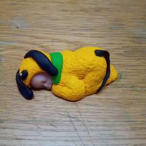 May include: A yellow clay figurine of a sleeping baby wearing a black and green dog costume.