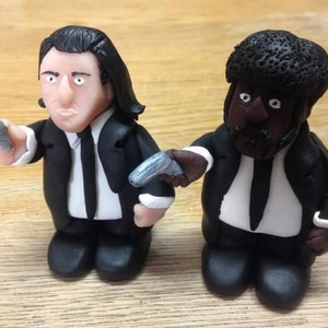 May include: Two handmade figurines of men in black suits, one with a black afro wig and the other with long black hair. Both figurines are holding toy guns.