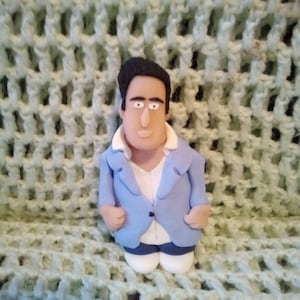 May include: A blue and white figurine of a person with black hair. The figurine is wearing a blue blazer, a white shirt, and blue trousers. The figurine is standing on a green crocheted surface.