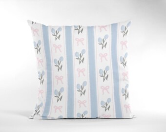 Puede incluir: Cojín cuadrado con un patrón repetido de rayas verticales azules, flores azul claro y lazos rosas sobre un fondo blanco. Un elemento decorativo para añadir un toque de elegancia a cualquier habitación.