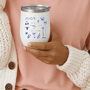 May include: A white stainless steel tumbler with a clear lid featuring blue line art illustrations of waves, a bikini, a seashell, palm trees, a cocktail glass, and the text "SUN, SEA & SELF-CARE" printed in a simple, thin, dark blue font.