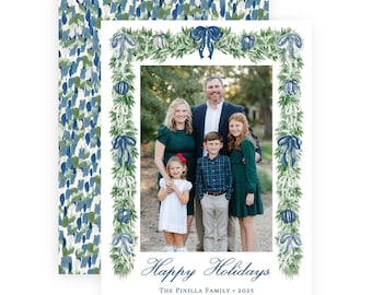 May include: A rectangular holiday card featuring a family photograph framed by a watercolor illustration of green pine garlands, blue bows, and blue ornaments. The text reads Happy Holidays, The Pinilla Family, 2025, and the names of the family members.