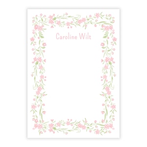 May include: A white card with a watercolor floral border of pink flowers and green leaves. The text "Caroline Wilt" is in the center of the card in a light pink color. The floral border frames the text.