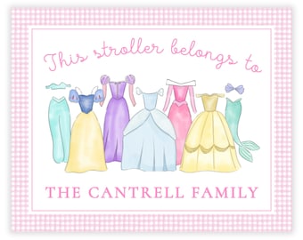 以下が含まれることがあります： ピンクと白のギンガムチェックの背景に、「This stroller belongs to THE CANTRELL FAMILY」というテキストと、ドレスとアクセサリーをつけた7人のプリンセスキャラクターが並んでいます。