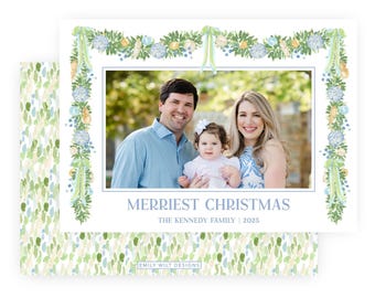 May include: A rectangular holiday greeting card featuring a central photograph of a family of three framed by a watercolor illustration of green garlands, blue and yellow flowers, and ribbons. The text reads MERRIEST CHRISTMAS THE KENNEDY FAMILY 2025.