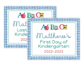 May include: Two blue and white gingham patterned signs with a white background. The top sign says "A B C Mat Last Kinde 202". The bottom sign says "A B C Matthere's First Day of Kindergarten 2022-2023".