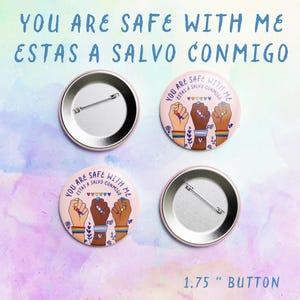 May include: Two 1.75 inch button pins with a pink background. The front of the button features a design of three fists raised in solidarity, with rainbow bracelets on each wrist. The text "You are safe with me" and "Estas a salvo conmigo" is written in a colorful font around the fists. The back of the button is plain silver.