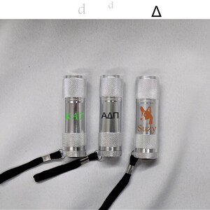 May include: Three silver flashlights with black straps. The flashlights are engraved with the text "KAT", "AΔΠ", and "Suzy" with a small dog silhouette.